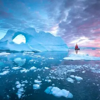 Little red sailboat cruising among floating icebergs in Disko Bay glacier during midnight sun season, Greenland